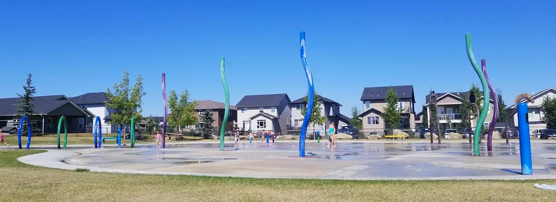 A splash park with colourful water sculptures in front of houses in High River